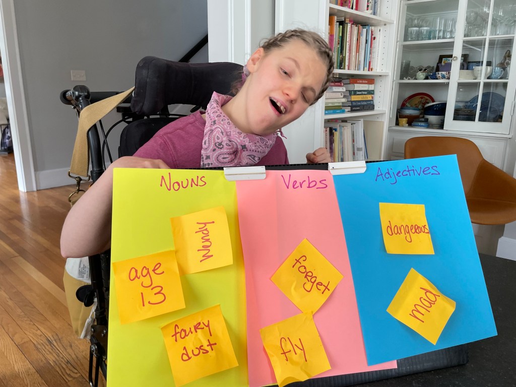 A student in a pink shirt sits in her wheelchair and smiles at the camera with a completed vocabulary sorting board activity in her lap.