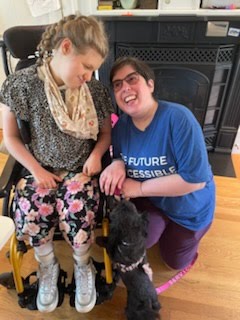 Teacher Sharon Rose kneels next to a student seated in her wheelchair. Student and teacher smile at each other.
